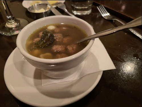 A bowl of soup with meatballs and greens, served on a white plate with a napkin, on a dark wooden table.