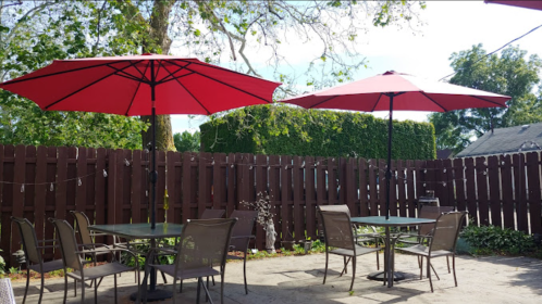 A patio with two red umbrellas over tables and chairs, surrounded by a wooden fence and greenery.