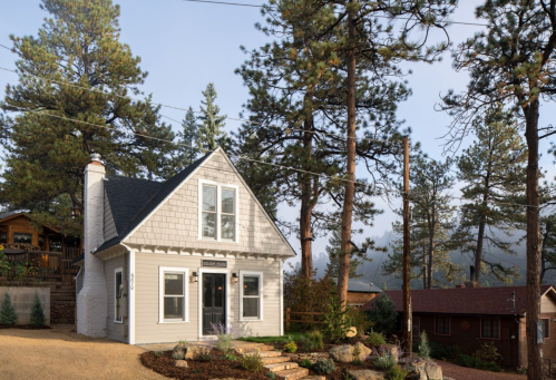 A charming two-story house surrounded by tall pine trees, with a gravel path leading to the entrance.
