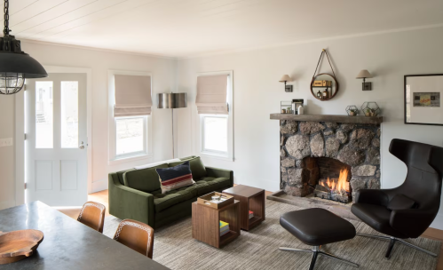 Cozy living room with a stone fireplace, green sofa, modern chair, and natural light from large windows.
