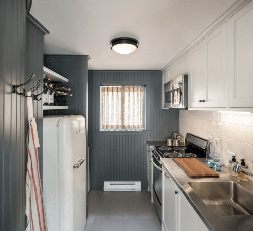 A modern kitchen with gray paneling, white cabinets, stainless steel appliances, and a window with blinds.