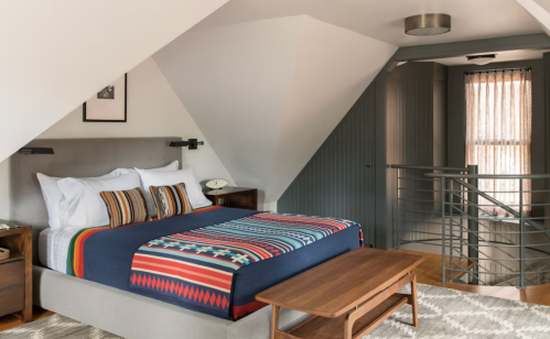 Cozy attic bedroom with a patterned blanket, wooden furniture, and large windows, featuring modern decor and natural light.