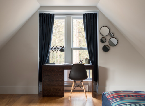 A cozy workspace with a desk, chair, lamp, and mirrors, framed by large windows with dark curtains.