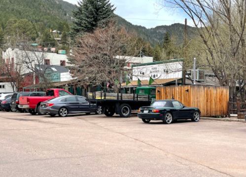 A parking lot with several cars, a trailer, and a backdrop of trees and buildings in a mountainous area.
