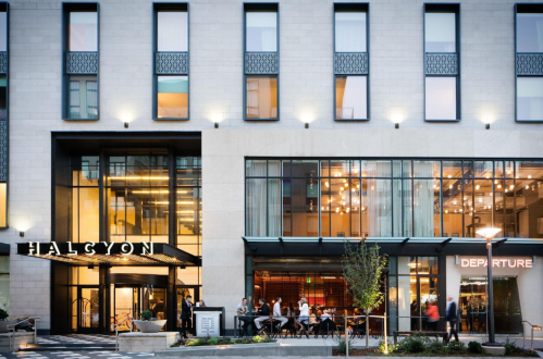 Exterior view of a modern building with the name "Halcyon" and outdoor seating area, featuring large windows and lights.