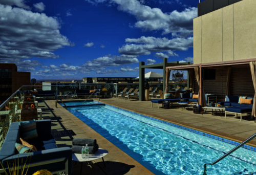 Rooftop pool area with lounge chairs, blue water, and a scenic sky filled with clouds.