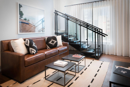 A modern living room featuring a brown leather sofa, patterned pillows, glass staircase, and minimalist decor.