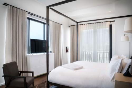 Modern bedroom with a four-poster bed, TV, and large windows draped with light curtains.