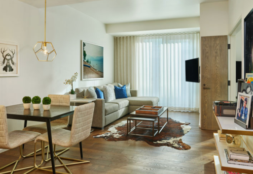 Modern living room with a gray sofa, blue cushions, dining table, and large windows with sheer curtains.