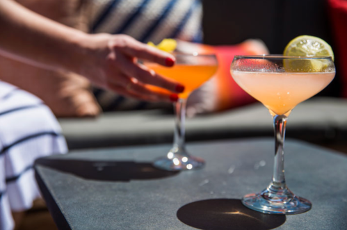 A hand reaches for a cocktail on a table, with two colorful drinks garnished with lime slices in the foreground.