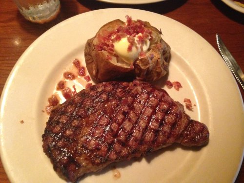 A grilled steak with a baked potato topped with sour cream and bacon bits on a white plate.