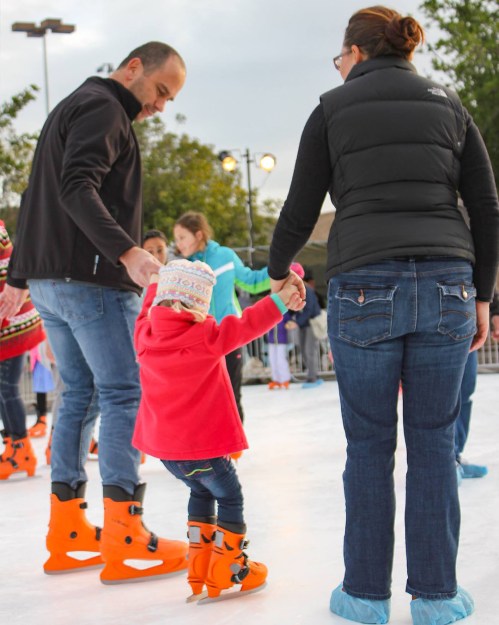 A man and woman help a young child skate on ice, all wearing skates and winter clothing, with others skating in the background.