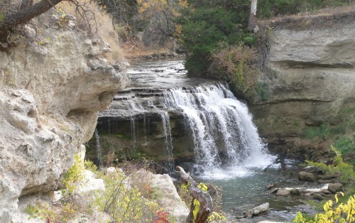 A serene waterfall cascades over rocky cliffs, surrounded by autumn foliage and a tranquil river.
