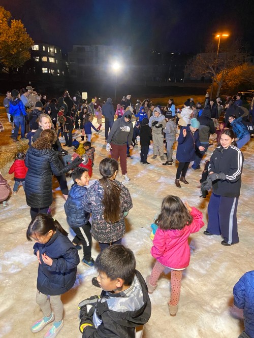 A lively outdoor event at night with many people, including children, playing and dancing on a snowy surface.