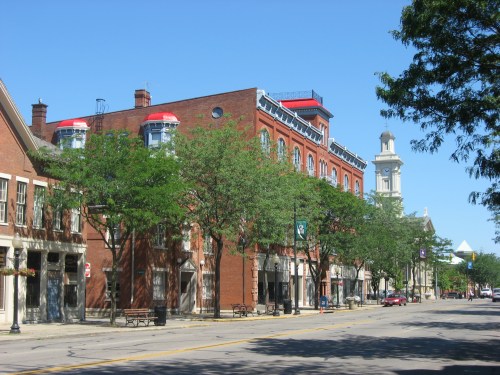 A sunny street view featuring brick buildings, trees, and a clock tower in the background.