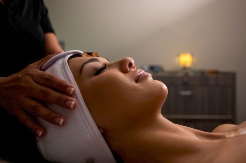 A person receiving a facial treatment, eyes closed, with a towel on their head and a hand gently massaging their forehead.