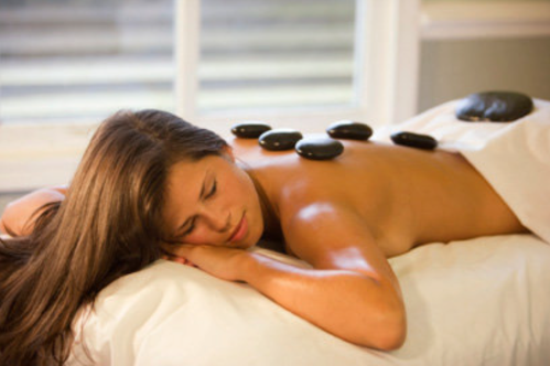 A woman lies on a massage table with hot stones on her back, eyes closed, enjoying a relaxing treatment.