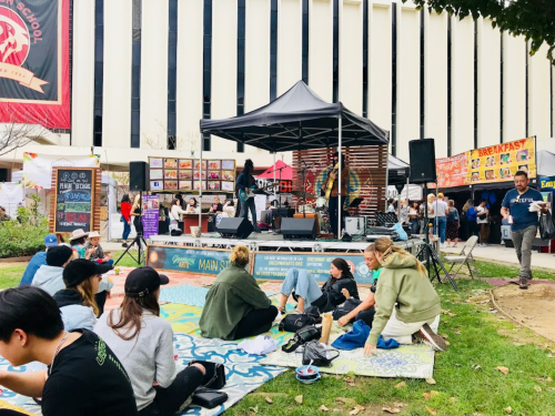 A lively outdoor event with a stage, food stalls, and people sitting on the grass enjoying the atmosphere.