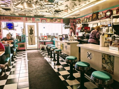 A retro diner with checkered floors, teal stools, and vintage decor, featuring patrons and a counter with a barista.
