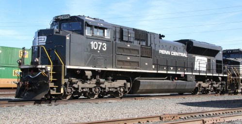 A black diesel locomotive with "PENNSYLVANIA CENTRAL" on the side, parked on railway tracks near freight containers.