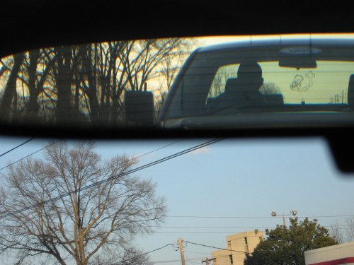 A view through a rearview mirror showing a car behind, with trees and a clear sky in the background.