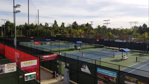 Aerial view of a tennis facility with multiple courts, players practicing, and surrounding greenery.