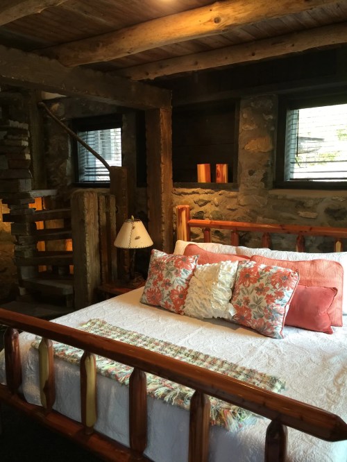 Cozy bedroom with a wooden bed, decorative pillows, stone walls, and a spiral staircase in a rustic setting.