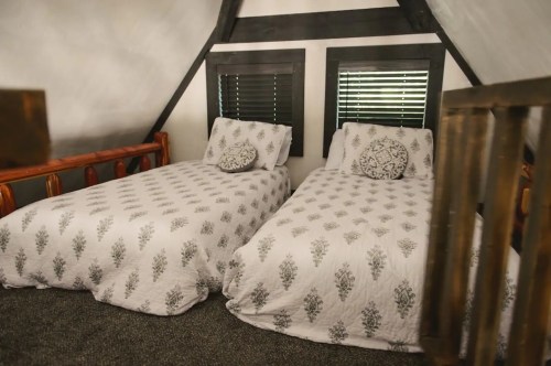 Cozy attic bedroom with two beds, patterned bedding, and windows with dark shutters. Warm, inviting atmosphere.
