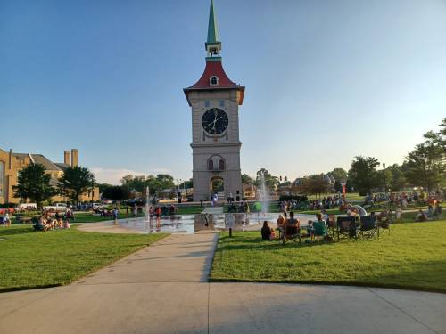 A clock tower stands in a park with people enjoying a splash pad and sitting on the grass under a clear blue sky.