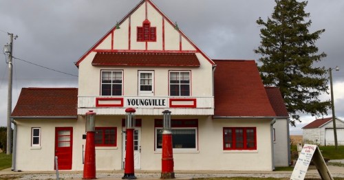 Historic building in Youngville with red accents and vintage gas pumps in front, set against a cloudy sky.