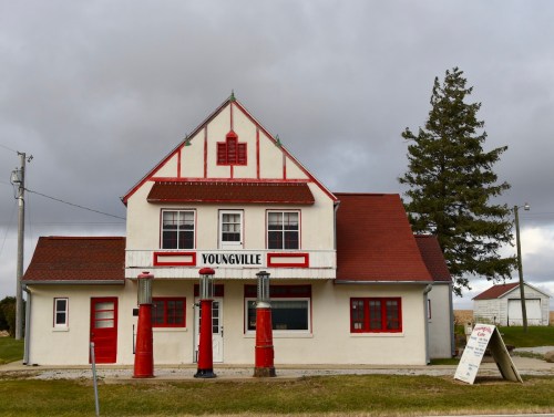 Historic building with red and white trim, featuring vintage gas pumps in front, set against a cloudy sky.