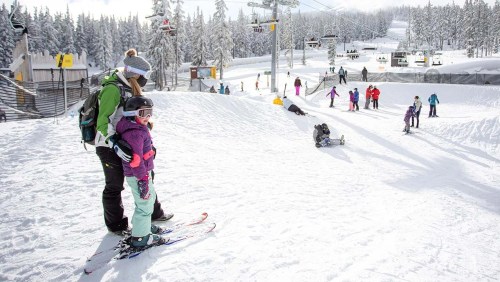 A parent and child in ski gear stand on a snowy slope, surrounded by skiers and a winter landscape.