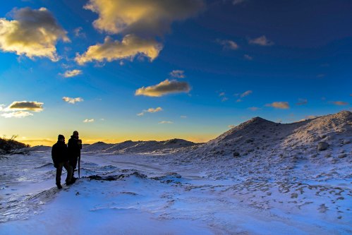 Two hikers stand on a snowy landscape, silhouetted against a vibrant sunset and a blue sky with scattered clouds.