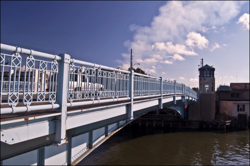 A blue bridge spans over water, with a control tower and buildings in the background under a partly cloudy sky.