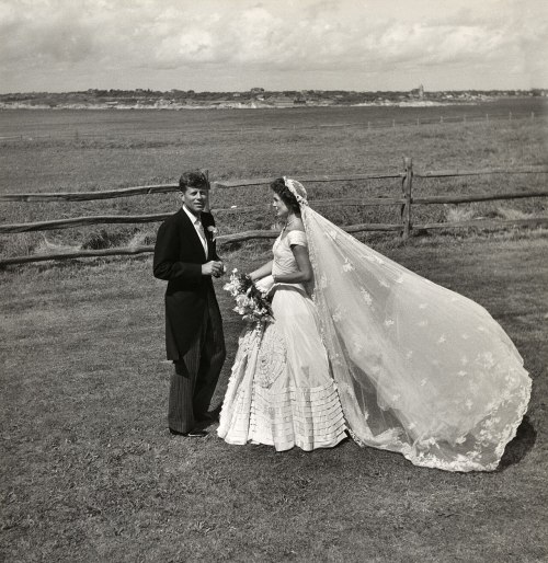 A couple in formal wedding attire stands outdoors, with a scenic landscape and a wooden fence in the background.