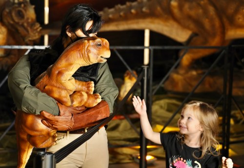 A person holds a dinosaur puppet while a smiling child waves, surrounded by dinosaur exhibits in a museum setting.