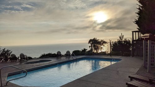 A serene poolside view at sunset, overlooking the ocean with lounge chairs and trees in the background.