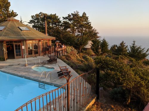 A scenic view of a poolside area with a hot tub, surrounded by trees and overlooking the ocean at sunset.