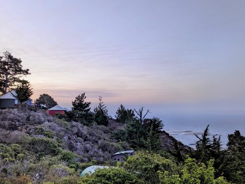 A coastal landscape at dusk, featuring hills, trees, and a view of the ocean under a soft, colorful sky.