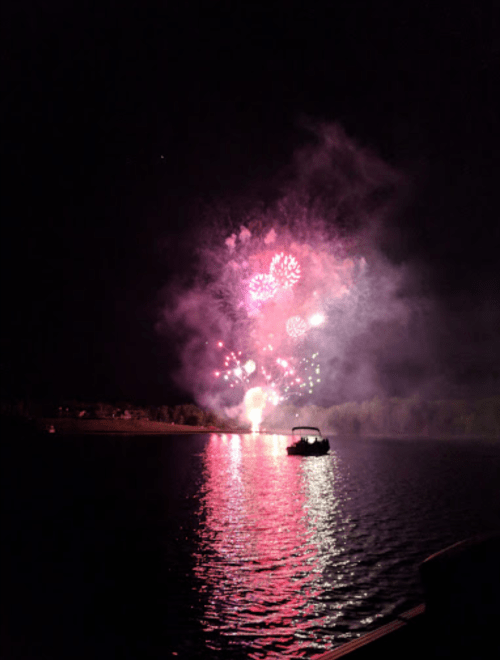 Colorful fireworks burst over a calm lake at night, reflecting vibrant hues on the water's surface.