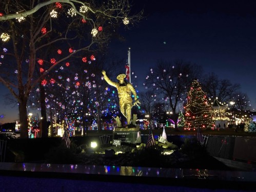 A golden statue of a soldier stands amidst colorful holiday lights and a decorated Christmas tree at dusk.
