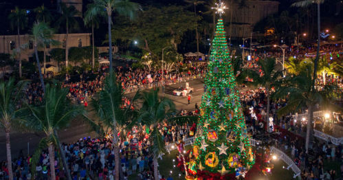 A festive Christmas tree surrounded by a large crowd at a holiday event, with palm trees and lights in the background.
