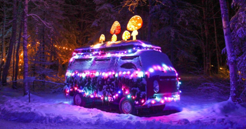 A colorful, illuminated van covered in snow, adorned with bright lights and mushroom decorations in a winter forest.