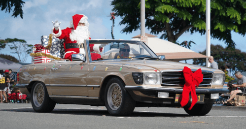 A convertible car decorated with a large bow, featuring Santa waving from the passenger seat during a parade.