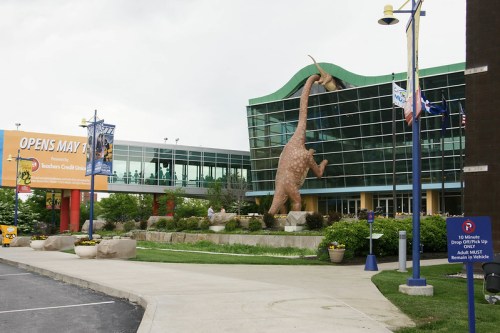 A large dinosaur statue stands outside a modern building with a sign announcing an opening date.