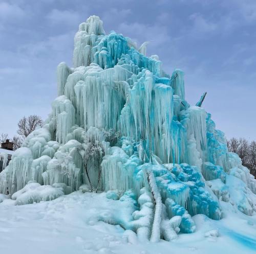 A towering ice formation with cascading blue and white icicles, surrounded by snow under a cloudy sky.