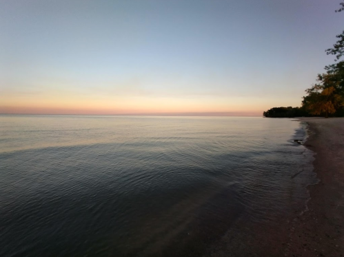 Serene beach at sunset, with calm waters reflecting soft pastel colors and a tree-lined shore in the distance.