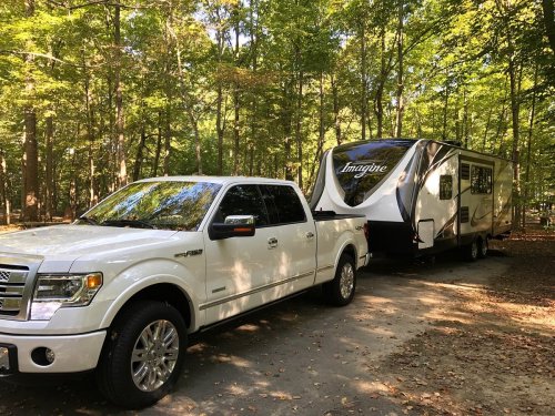 A white pickup truck towing a large travel trailer in a wooded area with green trees.