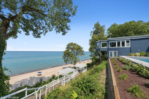 A scenic view of a beach with a house overlooking the water, surrounded by trees and greenery under a clear blue sky.