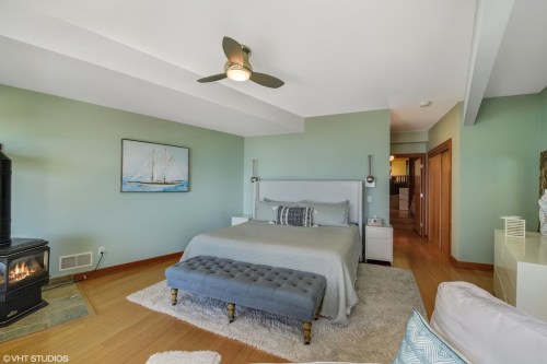 A cozy bedroom featuring a bed, blue ottoman, fireplace, and light green walls with a nautical painting.